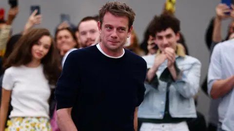 Reuters A man wearing a navy jumper and white top, he has dark hair