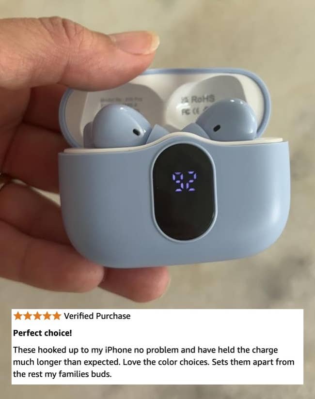 A hand holds a pair of wireless earbuds in an open charging case with a digital display showing the battery level