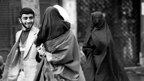 Getty Images A mullah in a white turban, holding his little daughter in black chador in his arms, talks to a man, while accompanied by his wife, also in black chador with a net mask covering her face, as they head towards Friday prayers at Tehran university, 1 February 1980