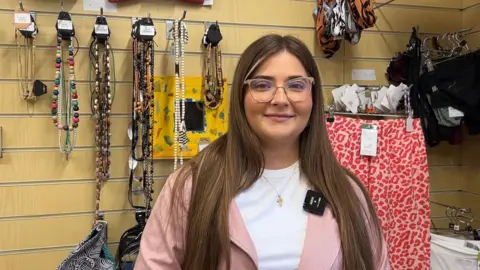 A woman in her twenties with long brown hair smiles down the camera. She is wearing a pale pink jacket and a white T-shirt, with beige rimmed glasses.