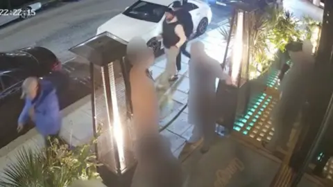PA Media A CCTV image of three men walking down a street past a building where a man is smoking outside