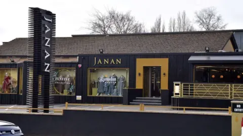 LDRS A black and gold single-storey building with display windows showing clothes. A large sign in the car park reads: Janan