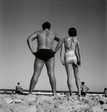 Black and white photograph of a man and a woman on a beach, wearing bathers, seen from behind