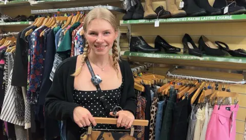 BBC A young woman with blonde hair in plaits smiles at the camera. She is standing in front of a rack of clothes inside a shop, holding a hanger with an item of clothing on it.