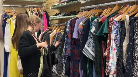 A young woman with blonde hair in plaits looks through rails of clothing in a shop, next to another woman who is facing away from the camera.