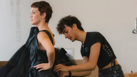 BBC Rohan Scott wearing a black T-shirt with laced up sides and black trousers, stands behind Lauren Oakley to adjust a dramatic black dress with a voluminous tulle skirt during a fitting session at Birmingham City University.